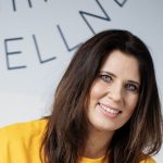 A woman with dark hair smiles in a yellow sweatshirt.