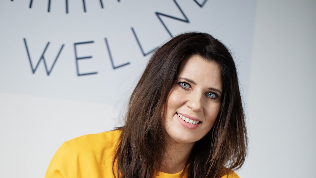 A woman with dark hair smiles in a yellow sweatshirt.